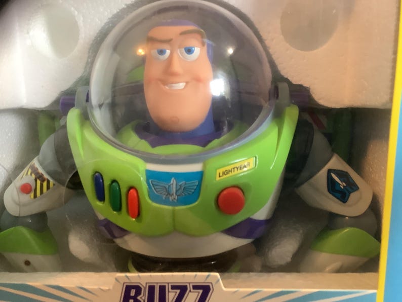 Op de afbeelding: Een Buzz Lightyear actiefiguur in de originele verpakking. De figuur heeft een groen en wit ruimtepak met een heldere helm, een lachend gezicht en het Lightyear-logo. De verpakking is geel en wit.
