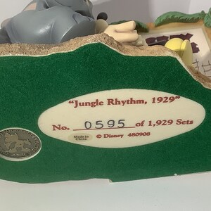 Puede incluir: Una base verde con una etiqueta ovalada blanca que dice "Jungle Rhythm, 1929" y "No. 0595 de 1.929 Sets". La etiqueta tambi&eacute;n incluye "Hecho en China" y "&copy; Disney 480908". La base presenta una escena esculpida de un elefante gris y un mono amarillo.