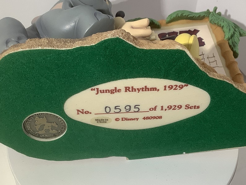 Puede incluir: Una base verde con una etiqueta ovalada blanca que dice "Jungle Rhythm, 1929" y "No. 0595 de 1.929 Sets". La etiqueta tambi&eacute;n incluye "Hecho en China" y "&copy; Disney 480908". La base presenta una escena esculpida de un elefante gris y un mono amarillo.