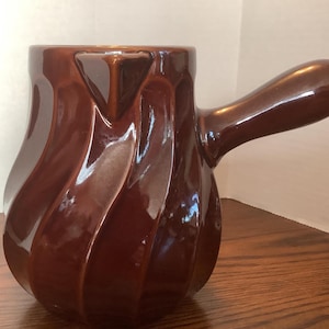 May include: A glossy, brown ceramic pitcher with a handle. The pitcher has a unique, swirled design and a spout for pouring. The handle is curved and attached to the side of the pitcher. The pitcher is likely used for serving beverages.
