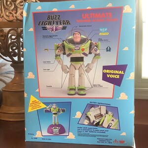 Op de afbeelding: Een boxed Buzz Lightyear Ultimate Talking Action Figure. De doos heeft een lichtblauwe achtergrond met wolken en illustraties van de kenmerken van het speelgoed, waaronder een intrekbare helm en een laserknop. De figuur is groen, wit en paars.