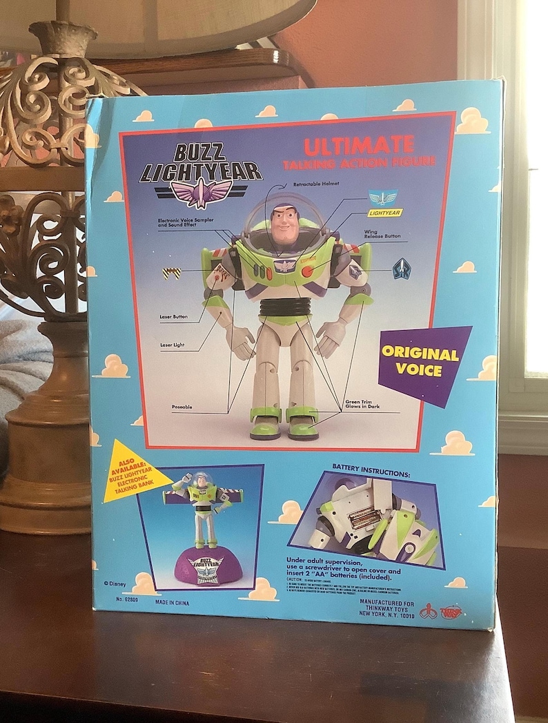 Op de afbeelding: Een boxed Buzz Lightyear Ultimate Talking Action Figure. De doos heeft een lichtblauwe achtergrond met wolken en illustraties van de kenmerken van het speelgoed, waaronder een intrekbare helm en een laserknop. De figuur is groen, wit en paars.