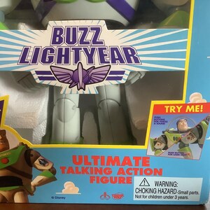 Op de afbeelding: Een verpakte Buzz Lightyear Ultimate Talking Action Figuur. De blauwe doos toont de tekst "Buzz Lightyear" en een afbeelding van het personage. De doos bevat ook de tekst "Original Voice" en "Try Me!"