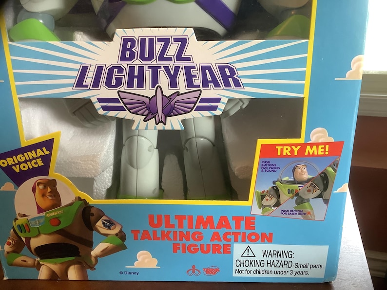 Op de afbeelding: Een verpakte Buzz Lightyear Ultimate Talking Action Figuur. De blauwe doos toont de tekst "Buzz Lightyear" en een afbeelding van het personage. De doos bevat ook de tekst "Original Voice" en "Try Me!"