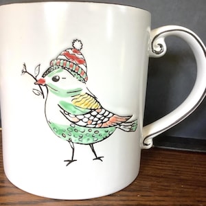 May include: White ceramic mug featuring a whimsical bird design. The bird is adorned with a striped hat and holds a sprig in its beak. The mug has a decorative handle with a silver trim.