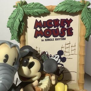 Puede incluir: Un conjunto de figuras que presenta a Mickey Mouse y un elefante gris. Mickey est&aacute; tocando un acorde&oacute;n y lleva una camisa roja y negra. El elefante est&aacute; de pie junto a Mickey. El conjunto est&aacute; delante de un cartel que dice "Mickey Mouse in Jungle Rhythm".