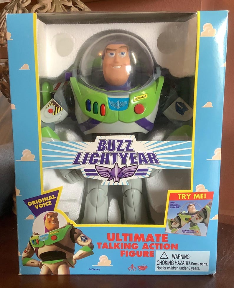 Op de afbeelding: Buzz Lightyear Ultimate Talking Action Figuur in een doos. De figuur zit in een doos met een blauwe lucht en wolken. De figuur draagt een groen en wit ruimtepak met een doorzichtige helm. Op de doos staan de woorden "Buzz Lightyear" en "Original Voice".
