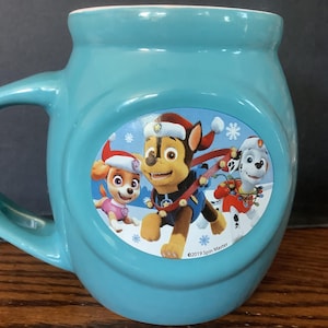 May include: A teal ceramic mug featuring a festive Paw Patrol design. The mug showcases Chase, Skye, and Marshall in Santa hats and winter attire. The mug has a curved handle and a white interior.