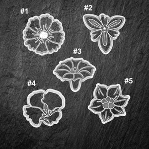 May include: Five white plastic cookie cutters shaped like flowers. The cutters are numbered 1 through 5.