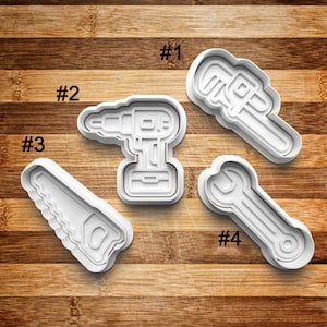 May include: Four white plastic cookie cutters shaped like tools: a wrench, a saw, a drill, and a pipe wrench. The cutters are numbered 1 through 4.