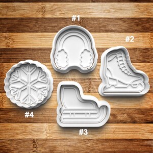 May include: Four white plastic cookie cutters shaped like a snowflake, a pair of earmuffs, a skate, and a skate with a sled. The cutters are numbered 1 through 4.