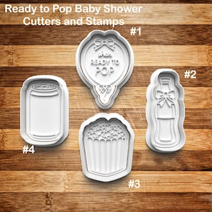 May include: A set of white plastic cookie cutters and stamps for a baby shower. The shapes include a balloon with the text "she's READY TO POP", a soda bottle, a popcorn container, and a soda can. The cutters are numbered 1 through 4.