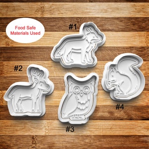 May include: Four white plastic cookie cutters shaped like a fox, deer, owl, and squirrel. The cutters are numbered 1 through 4. The text "Food Safe Materials Used" is visible in the top left corner of the image.