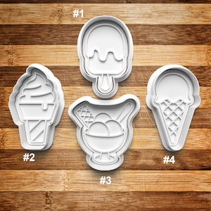 May include: Four white plastic cookie cutters shaped like different ice cream treats: a popsicle, a cone with soft serve, a sundae, and a cone with a scoop of ice cream. The cutters are numbered 1 through 4.