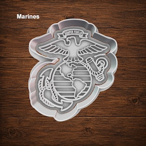 May include: A light gray cookie cutter shaped like the United States Marine Corps emblem, featuring an eagle, a globe, and an anchor. The design includes a banner with the text Semper Fidelis at the top and the word Marines in the corner.