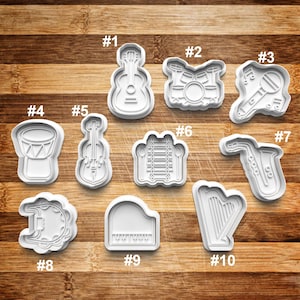 May include: Ten white plastic cookie cutters shaped like musical instruments, including a guitar, drums, a saxophone, a xylophone, a violin, a piano, a tambourine, a harp, a drum, and a clarinet. The cutters are numbered 1 through 10.