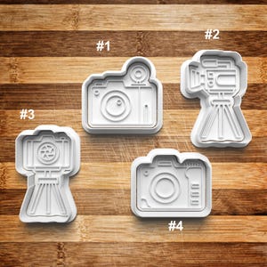 May include: Four white plastic cookie cutters shaped like cameras. The cutters are numbered 1 through 4. The cutters are on a wooden surface.