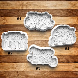 May include: Four white plastic cookie cutters with the words "Merry Christmas" in various fonts. The cutters are on a wooden surface. Each cutter has a different design, including script and block lettering.