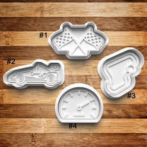 May include: Four white plastic cookie cutters shaped like a race car, a checkered flag, a racetrack, and a speedometer. The cutters are numbered 1 through 4.