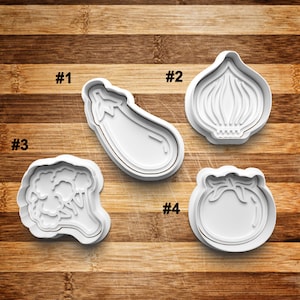 May include: Four white plastic cookie cutters shaped like vegetables: an eggplant, an onion, a broccoli, and a tomato. The cutters are numbered 1 through 4.