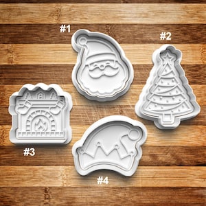 May include: Four white plastic Christmas cookie cutters. The cutters are shaped like Santa Claus, a Christmas tree, a fireplace, and a Santa hat. The cutters are numbered 1 through 4.