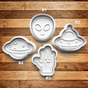 May include: Five white plastic cookie cutters shaped like an alien head, a flying saucer, a hand, a planet, and a ring around a planet. The cutters are numbered 1 through 4.