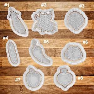 May include: Eight white plastic cookie cutters shaped like different vegetables, including a pea pod, green beans, lettuce, cucumber, chili pepper, cabbage, bell pepper, and a turnip.