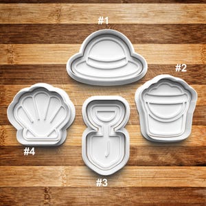 May include: Set of five white plastic cookie cutters shaped like a sun hat, seashell, bucket, shovel, and a sandcastle. The cutters are numbered 1 through 5.