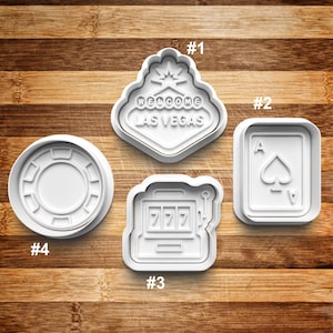 May include: Four white plastic cookie cutters shaped like a casino chip, a playing card, a slot machine, and a welcome to Las Vegas sign.