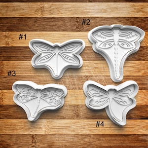May include: Four white plastic cookie cutters shaped like dragonflies. The cutters are numbered 1 through 4.