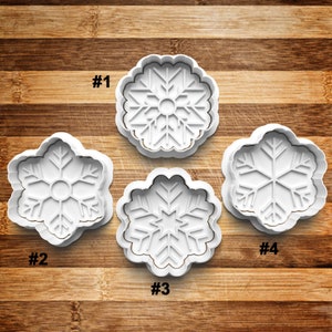 May include: Four white plastic snowflake cookie cutters on a wooden surface. The cutters are numbered 1 through 4.