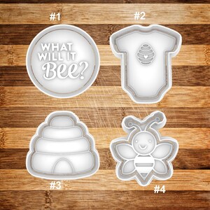 May include: Four cookie cutters in white plastic, shaped like a circle with the text "What will it bee?", a baby onesie with a honeycomb design, a beehive, and a bee.