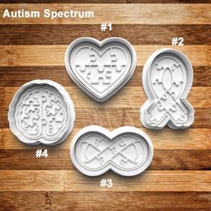 May include: Four white cookie cutters with puzzle piece designs. The shapes include a heart, a ribbon, an infinity symbol, and a brain. The text "Autism Spectrum" is at the top, with numbers labeling each cutter.