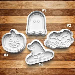 May include: Four white plastic Halloween-themed cookie cutters. The shapes include a ghost, a cluster of bats, a jack-o'-lantern, and a witch's hat. The cutters are arranged on a wooden surface.