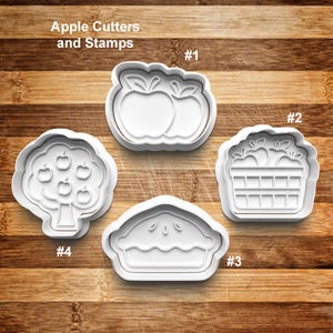 May include: Four white plastic apple-themed cookie cutters and stamps on a wooden surface. The cutters include two apples, an apple basket, an apple pie, and an apple tree. The text "Apple Cutters and Stamps" is at the top.