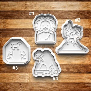 May include: Four white plastic cookie cutters, each with a different design. The first cutter is shaped like a person with a beard and wild hair. The second cutter is shaped like a volcano with a palm tree on top. The third cutter is shaped like a dog standing on a rock with two people on either side. The fourth cutter is shaped like a cave with a person holding a club and a fire in front of the cave.