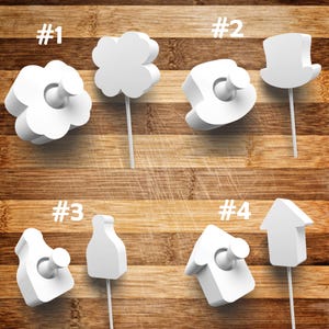May include: A collection of four white bottle stoppers in various shapes: a flower, a four-leaf clover, a top hat, and a house. Each stopper has a white stem and a circular stopper. The stoppers are displayed on a wooden surface.