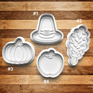 May include: Four white plastic cookie cutters, numbered 1 through 4, on a wooden surface. The cutters are shaped like a pilgrim hat, an oak leaf, a pumpkin, and an apple.