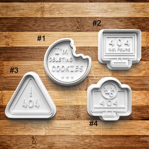 May include: Four white plastic cookie cutters shaped like a computer screen, a warning sign, a bitten cookie, and a computer screen with a sad face. The computer screens have the number 404 on them. The warning sign has an exclamation point and the number 404 on it. The cookie cutter shaped like a bitten cookie has the text "I'M DELETING COOKIES" on it.