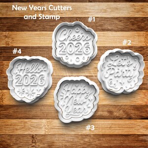 May include: Four white plastic cookie cutters with New Year's greetings. The cutters include the phrases "Cheers 2026", "Let's Party", "Happy New Year", and "Hello 2026". The cutters are on a wooden surface.