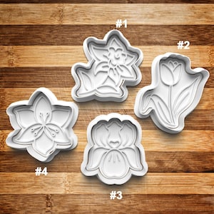 May include: Four white plastic cookie cutters shaped like flowers. The cutters are numbered 1 through 4. The flowers are a daffodil, a tulip, an iris, and a cherry blossom.