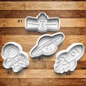May include: Four white plastic cookie cutters shaped like a satellite, astronaut, planet with rings, and a rocket. The cutters are numbered 1 through 4.