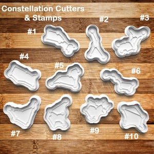 May include: A collection of ten white plastic constellation cookie cutters and stamps, each labeled with a number from one to ten. The cutters are arranged on a wooden surface, with the text "Constellation Cutters & Stamps" at the top.