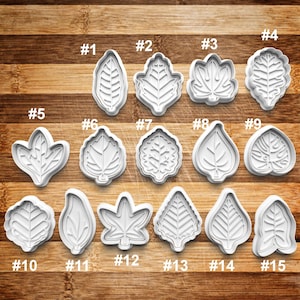May include: A collection of fifteen white plastic leaf-shaped cookie cutters, each with a unique design and labeled with numbers from #1 to #15. The cutters are arranged on a wooden surface, showcasing various leaf shapes and vein details.