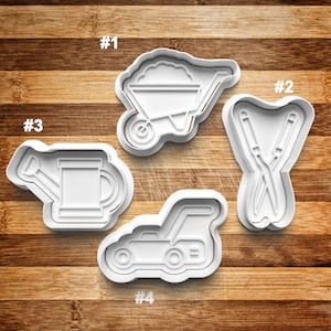 May include: Four white plastic cookie cutters shaped like gardening tools: a wheelbarrow, a pair of shears, a watering can, and a lawnmower. The cutters are numbered 1 through 4.