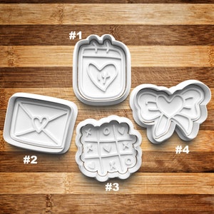 May include: Four white plastic cookie cutters with Valentine's Day themes. The cutters are numbered 1 through 4. Cutter #1 is a calendar with a heart inside. Cutter #2 is an envelope with a heart inside. Cutter #3 is a tic-tac-toe board with hearts and circles. Cutter #4 is a bow with a heart in the center.