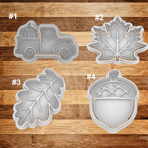 May include: Four different cookie cutters, numbered 1 through 4, on a wooden surface. The cutters are shaped like a pickup truck with a pumpkin, a maple leaf, an oak leaf, and an acorn.