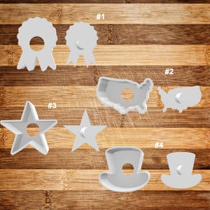 May include: Ten white plastic cookie cutters in various shapes, including a star, a ribbon, a hat, and the outline of the United States. The cutters are arranged on a wooden surface. The numbers 1, 2, 3, and 4 are written on the surface next to the cutters.