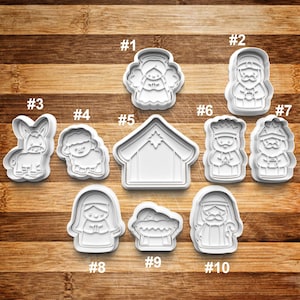 May include: A collection of white plastic cookie cutters in various shapes, including an angel, a shepherd, a donkey, a sheep, a stable, and figures of the Nativity. The cutters are arranged on a wooden surface.