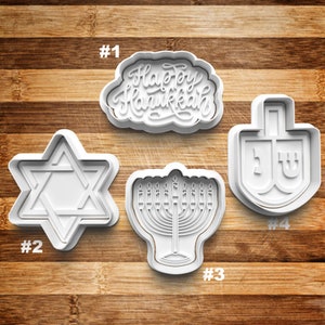 May include: Four white plastic cookie cutters for Hanukkah. The cutters are shaped like a Star of David, a menorah, a dreidel, and a banner that says "Happy Hanukkah".